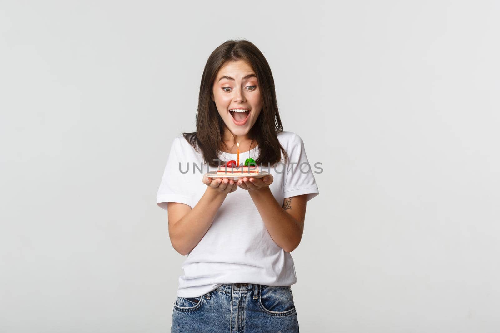 Excited attractive brunette b-day girl making wish on birthday cake, white background.