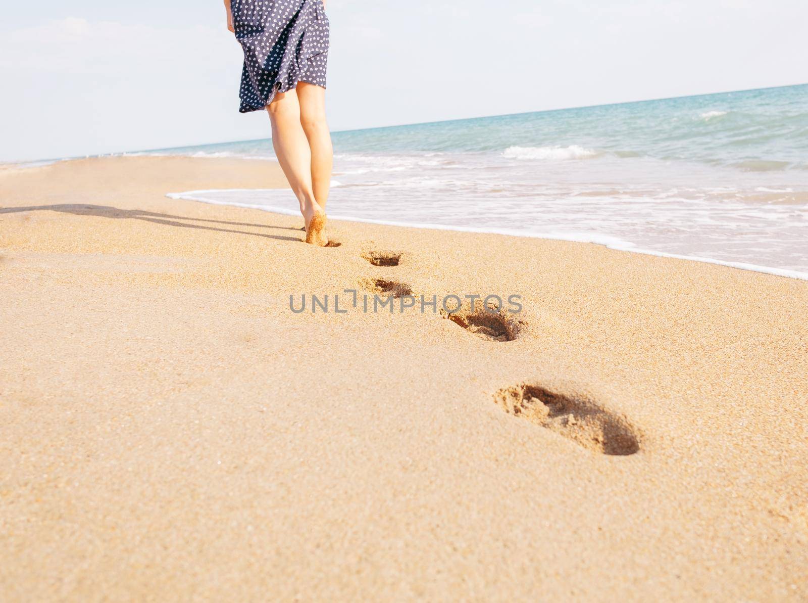 Barefoot young woman walking on beach leaving footprints in the sand near sea, view of legs. Concept of summer vacations.
