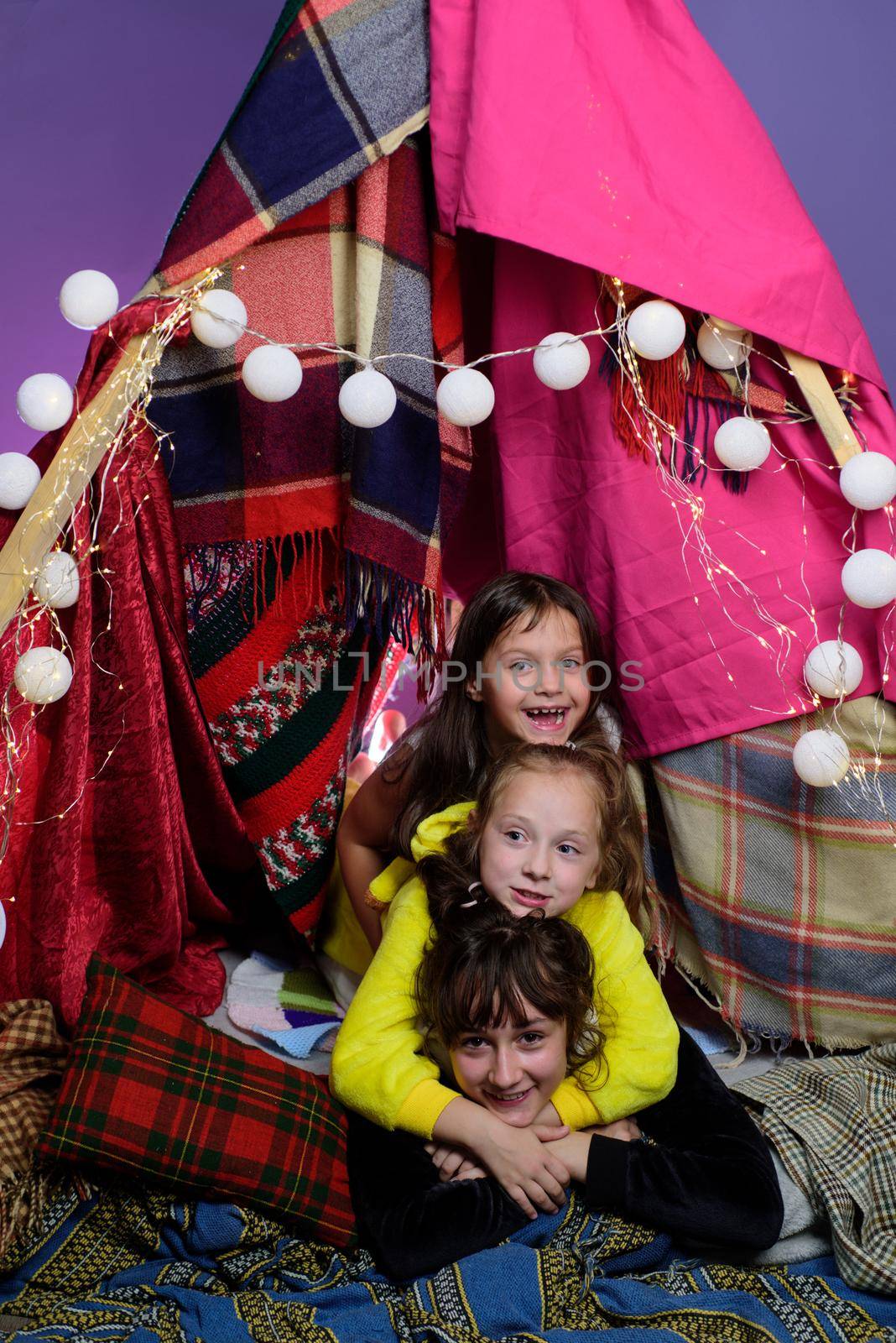 Kids embrace in home-made tent inside the living room before going to sleep. Child pajama party. Pajamas for kids. Little Girls in tipi house. Sisters or best friends spend time