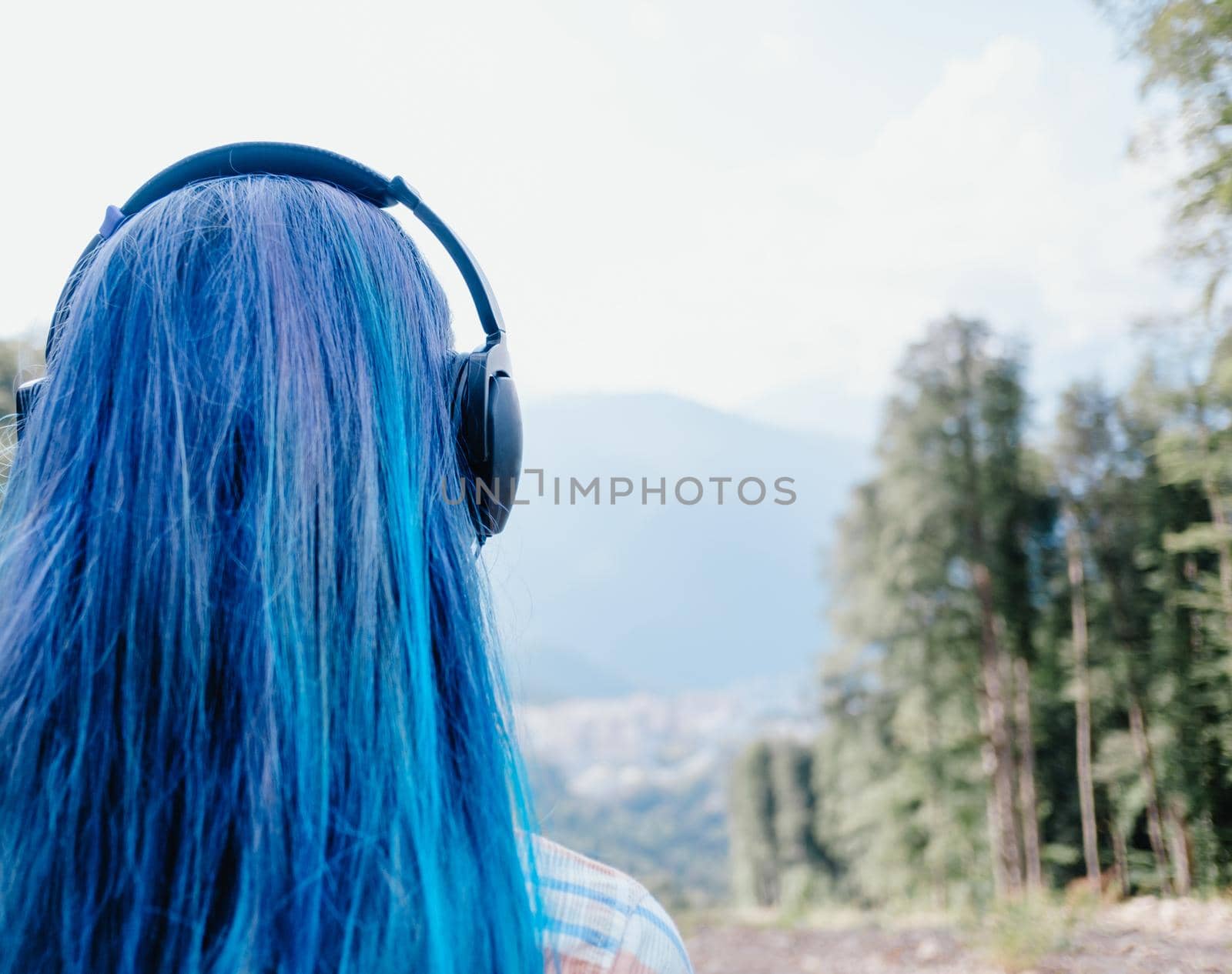 Girl with blue hair in headphones looking at summer landscape, rear view.