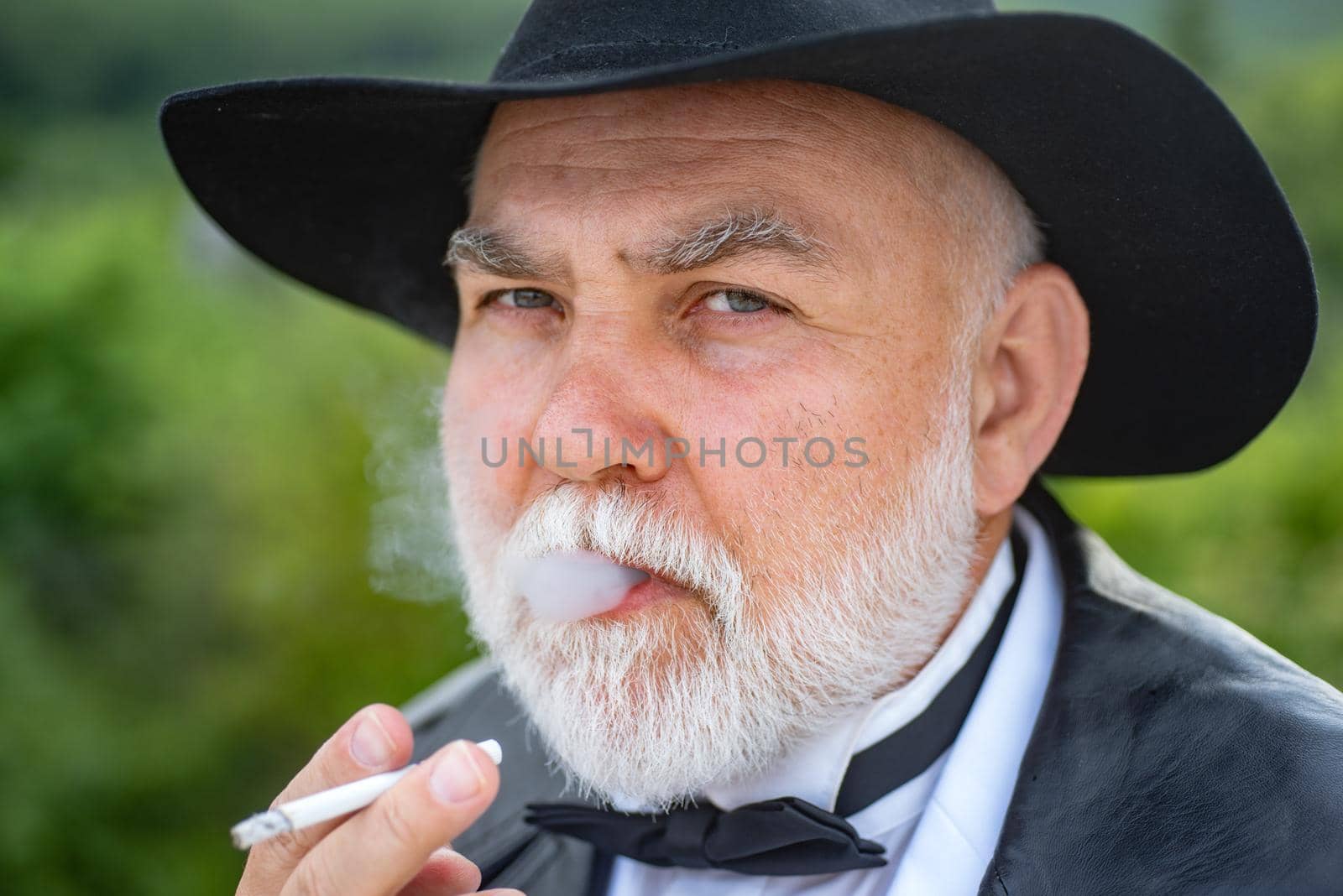 Close up face portrait of handsome senior with gray beard smoking cigarette. Attractive elderly mature man. Senior glamour vintage man wearing suit and tie and hat. Gangster look, Mafia pimp