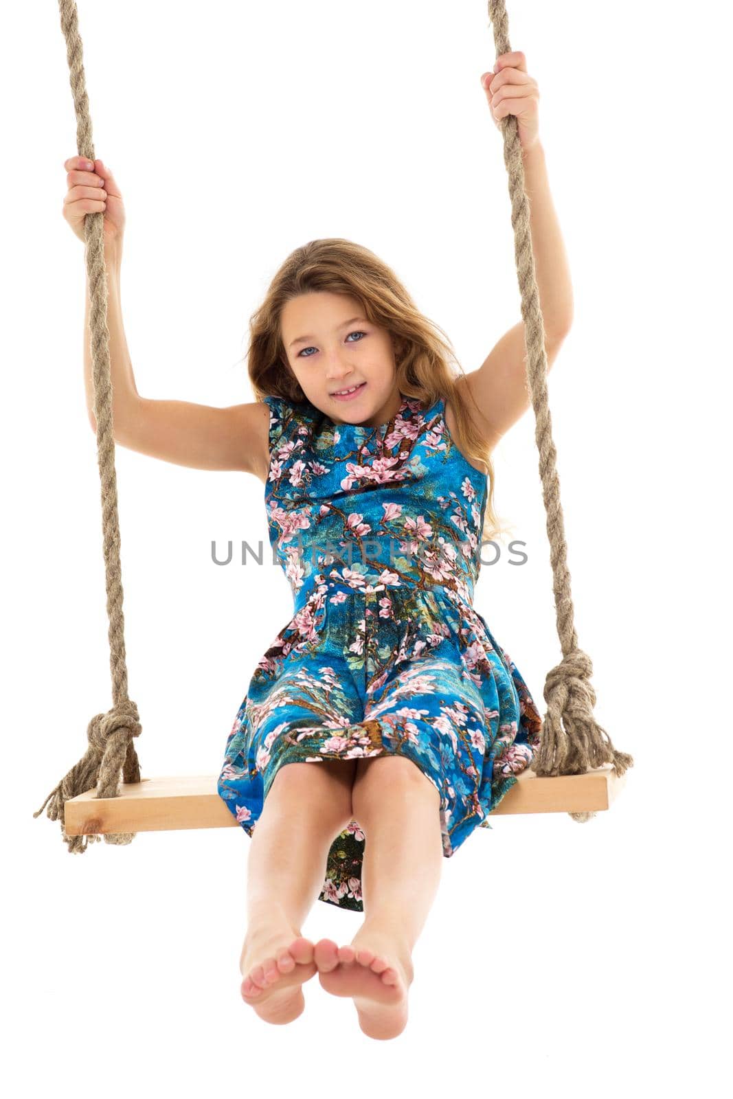 Blonde girl swinging on rope swing. Full length shot of pretty stylish barefoot girl having fun against white background. Portrait of preteen child posing in studio