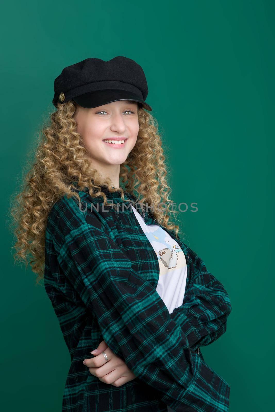 Portrait of happy beautiful girl in trendy outfit. Blonde girl with wavy hairstyle smiling at camera. Teenager wearing dark plaid cotton shirt and black cap posing on blue green background