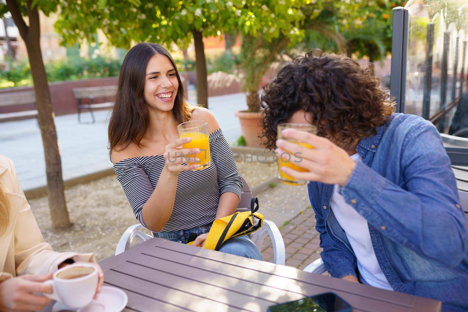 Couple of friends toasting with orange juices while having a drink with their multi-ethnic group of friends at the outdoor table of a bar.