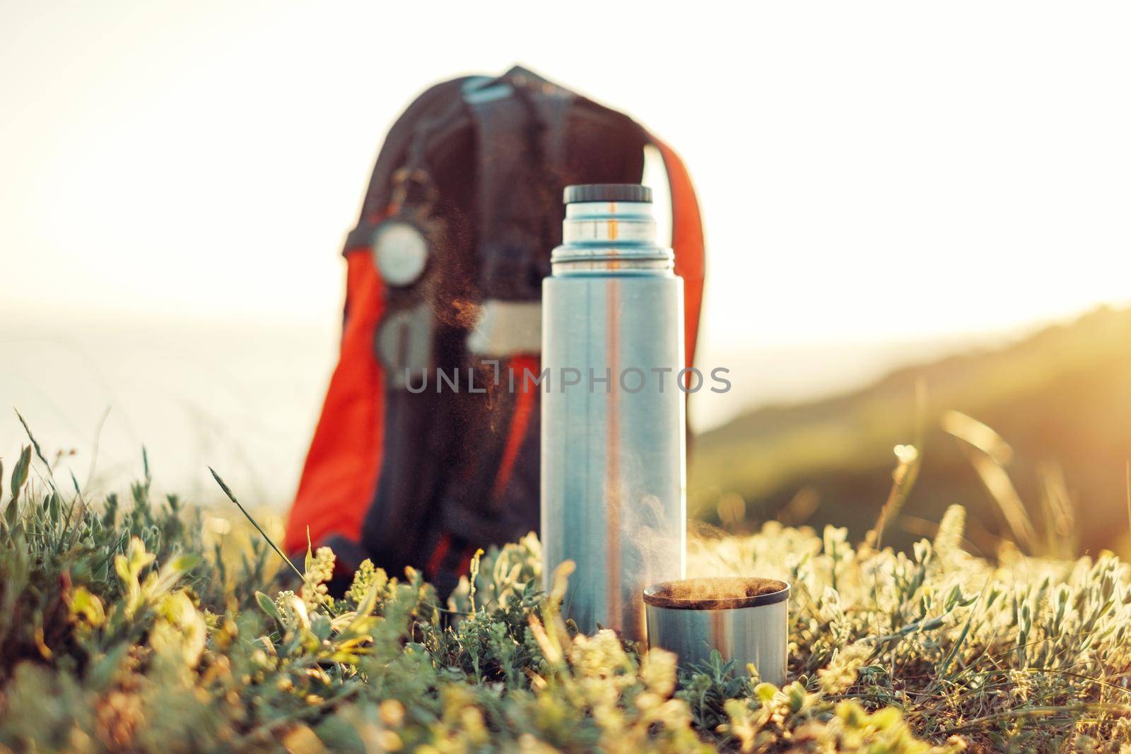Thermos and cup of hot drink near the backpack outdoor in summer.