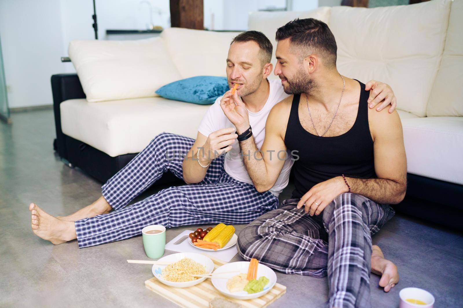 Gay couple eating together sitting on their living room floor. Homosexual lifestyle concept.