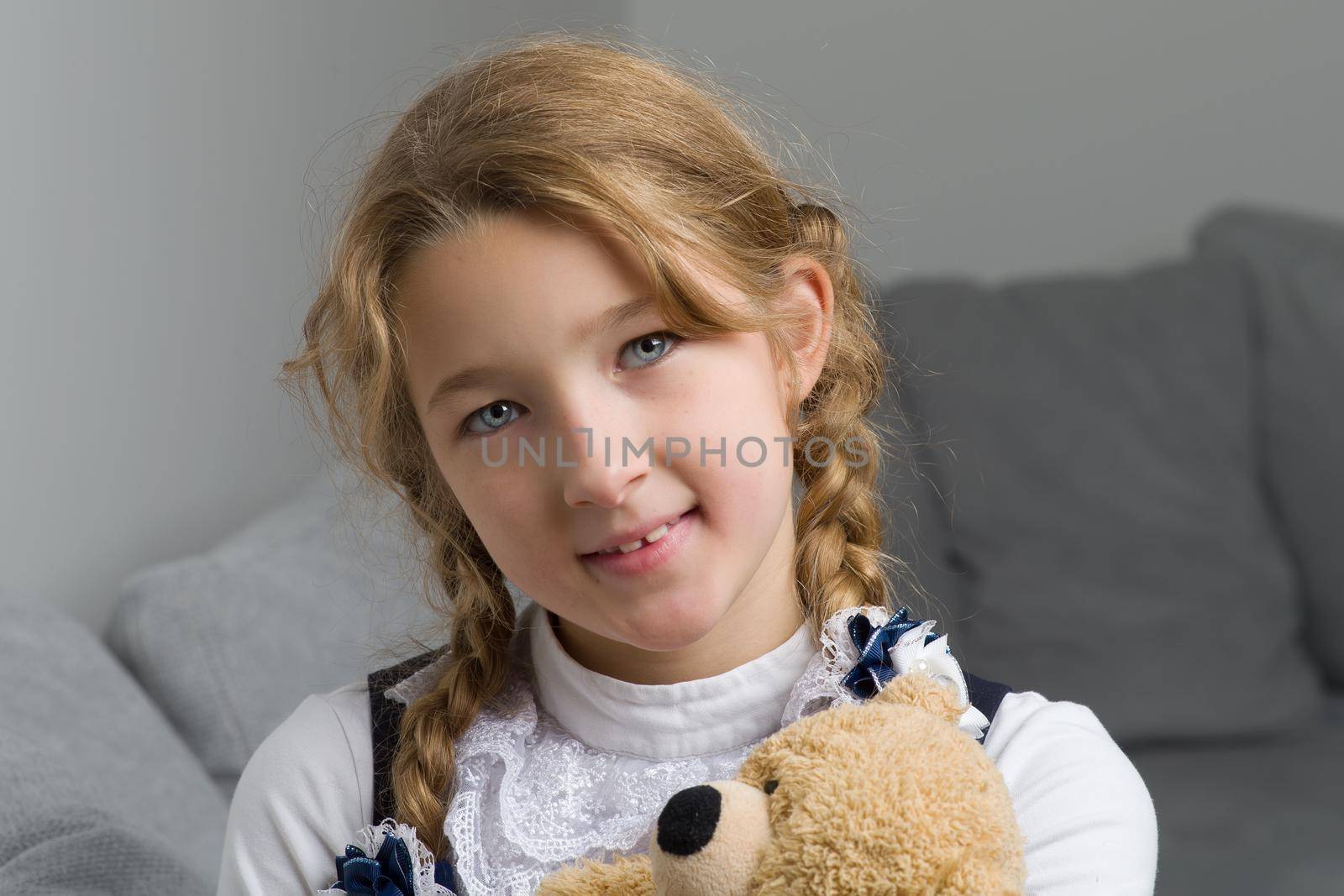 Cute schoolgirl sitting on sofa with teddy bear. Adorable primary school girl with braids dressed in uniform hugging her favorite toy. Back to school, education concept