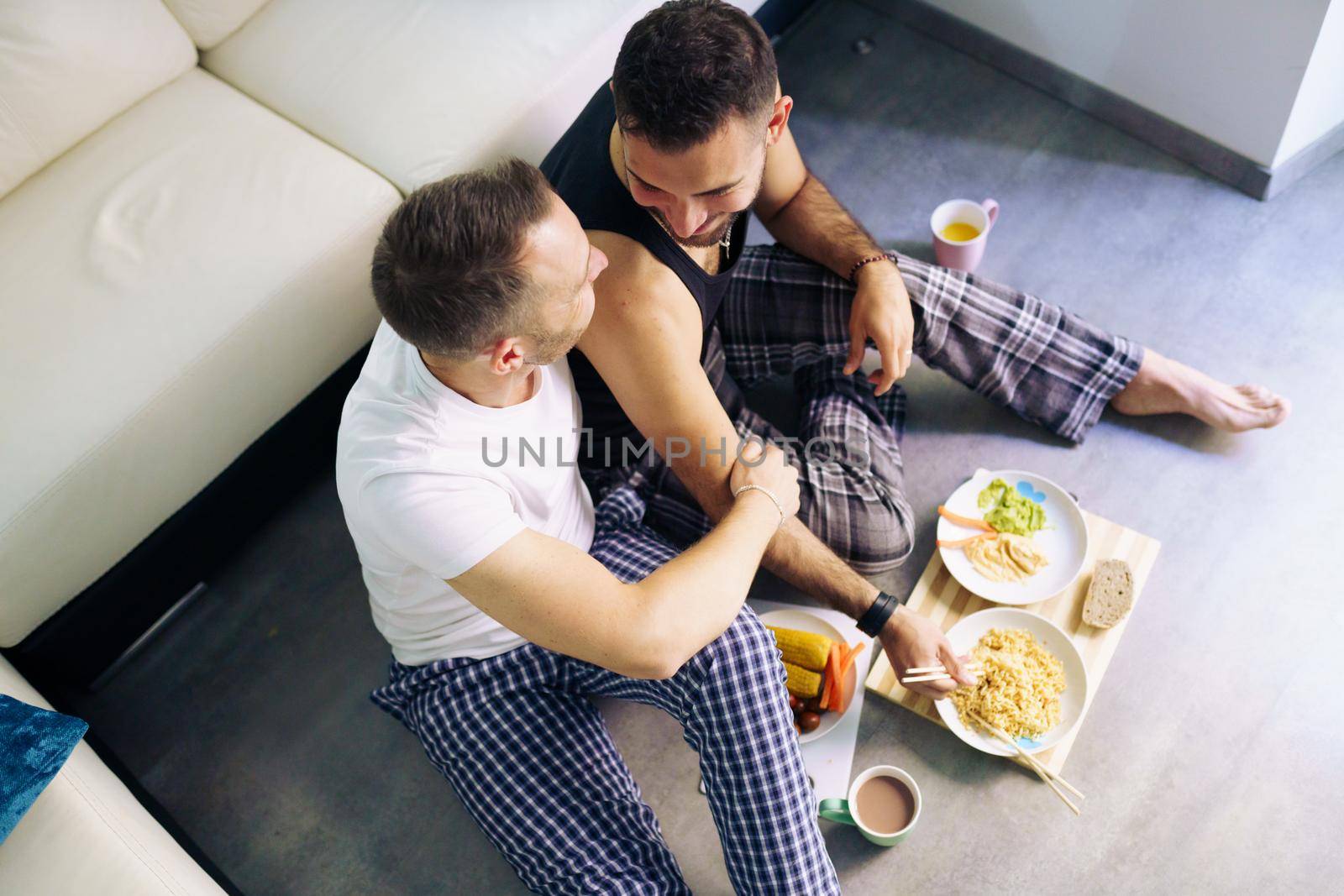 Gay couple eating together sitting on their living room floor. Homosexual lifestyle concept.