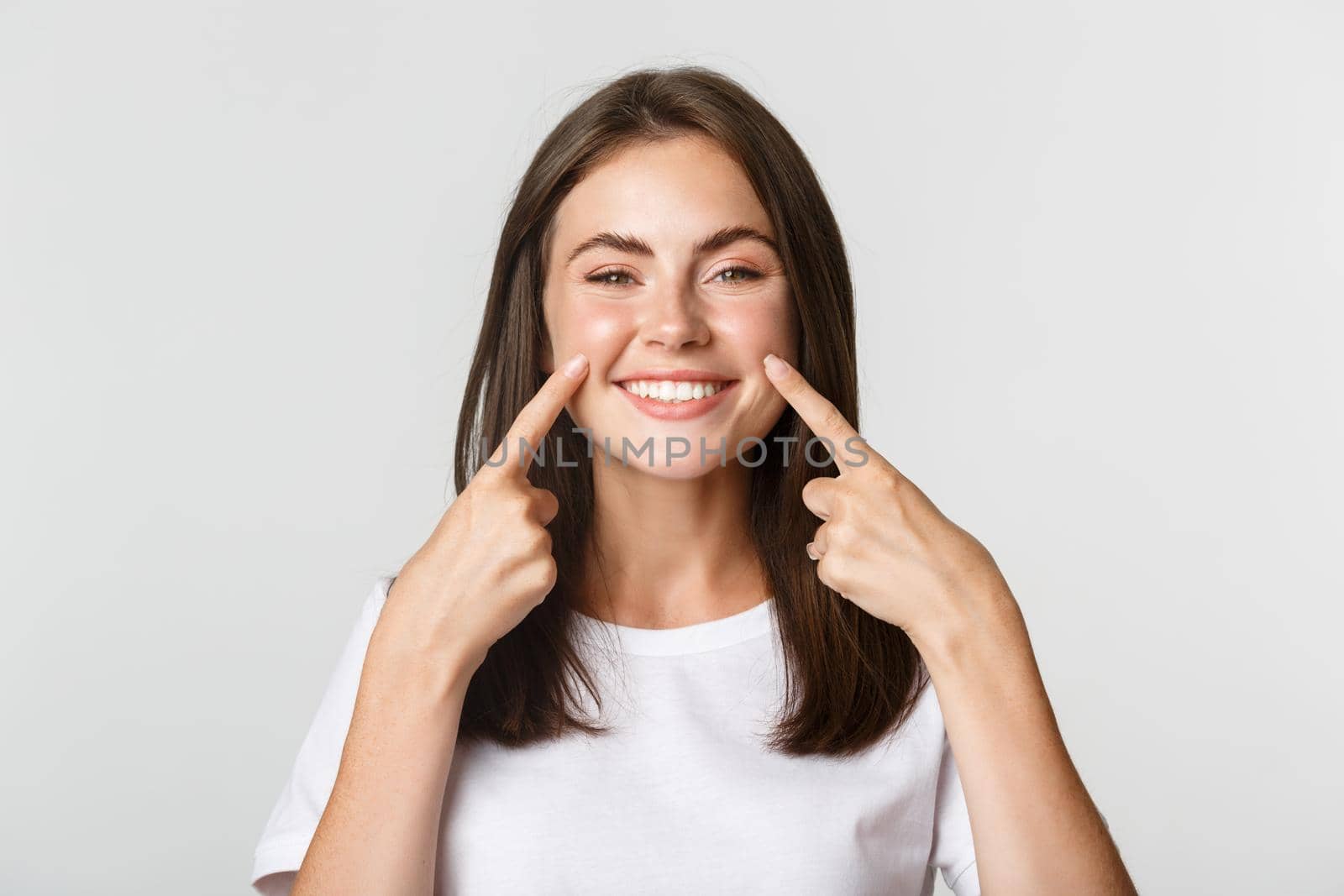 Close-up of attractive smiling young girl pointing fingers at face, poking cheeks, white background.