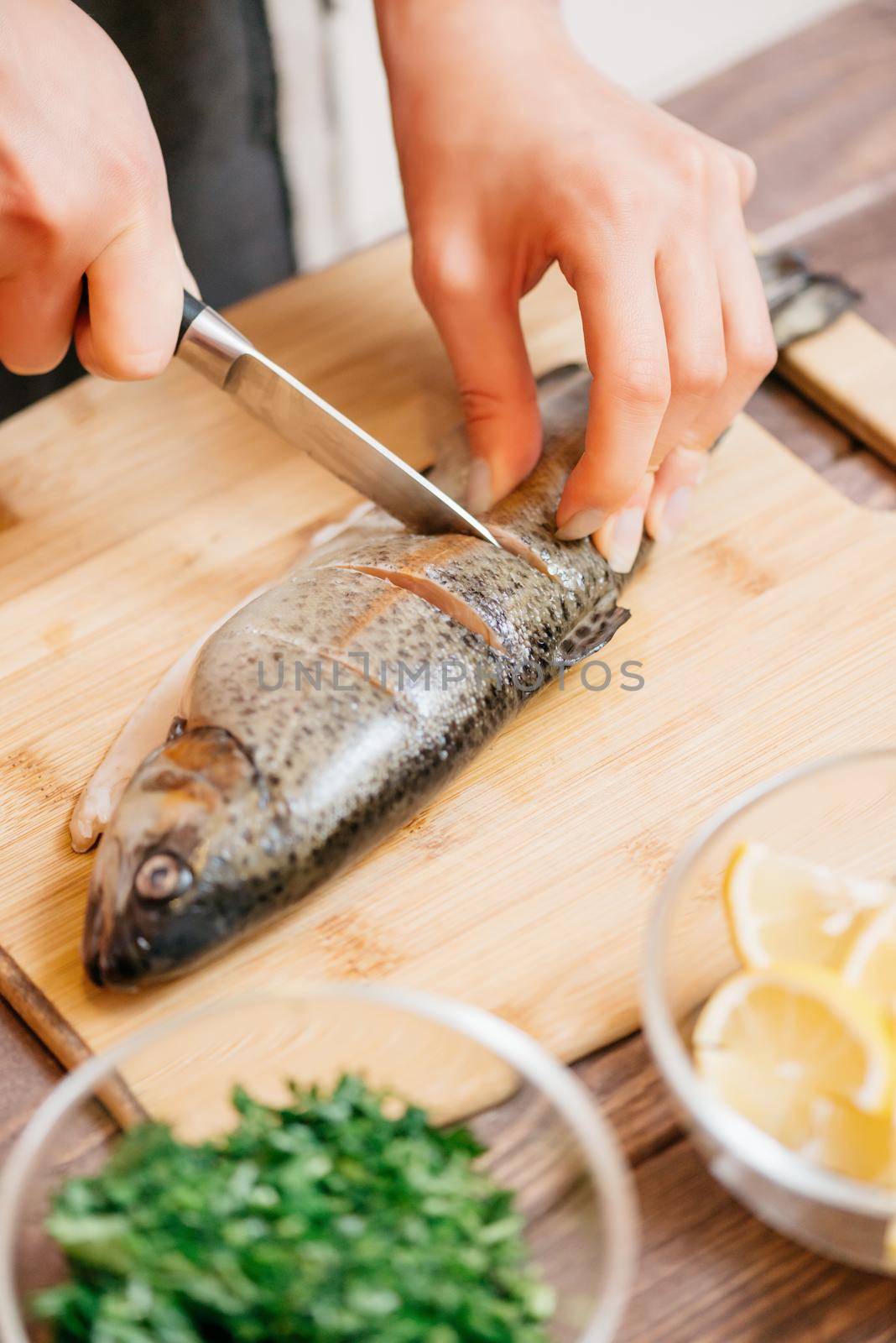 Woman cutting raw trout fish on wooden board for seafood dish in the kitchen, view of hands.