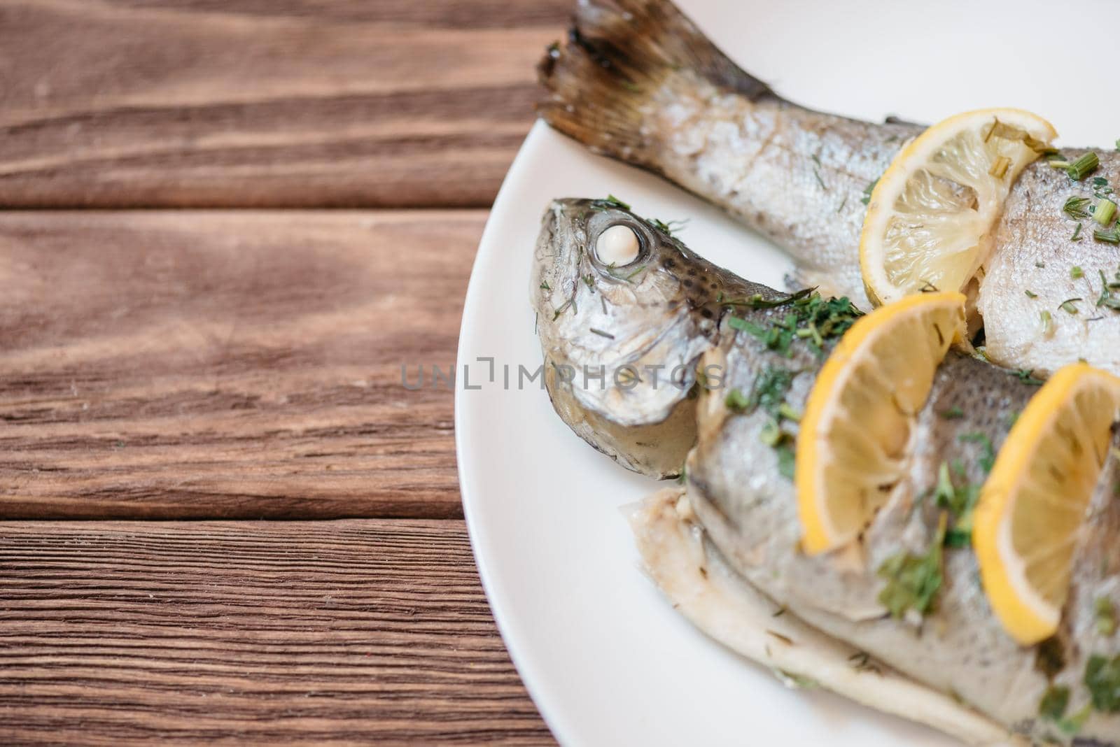 Simple fish dish with lemons and greenery on a white plate on wooden table. Copy-space in left part of image.