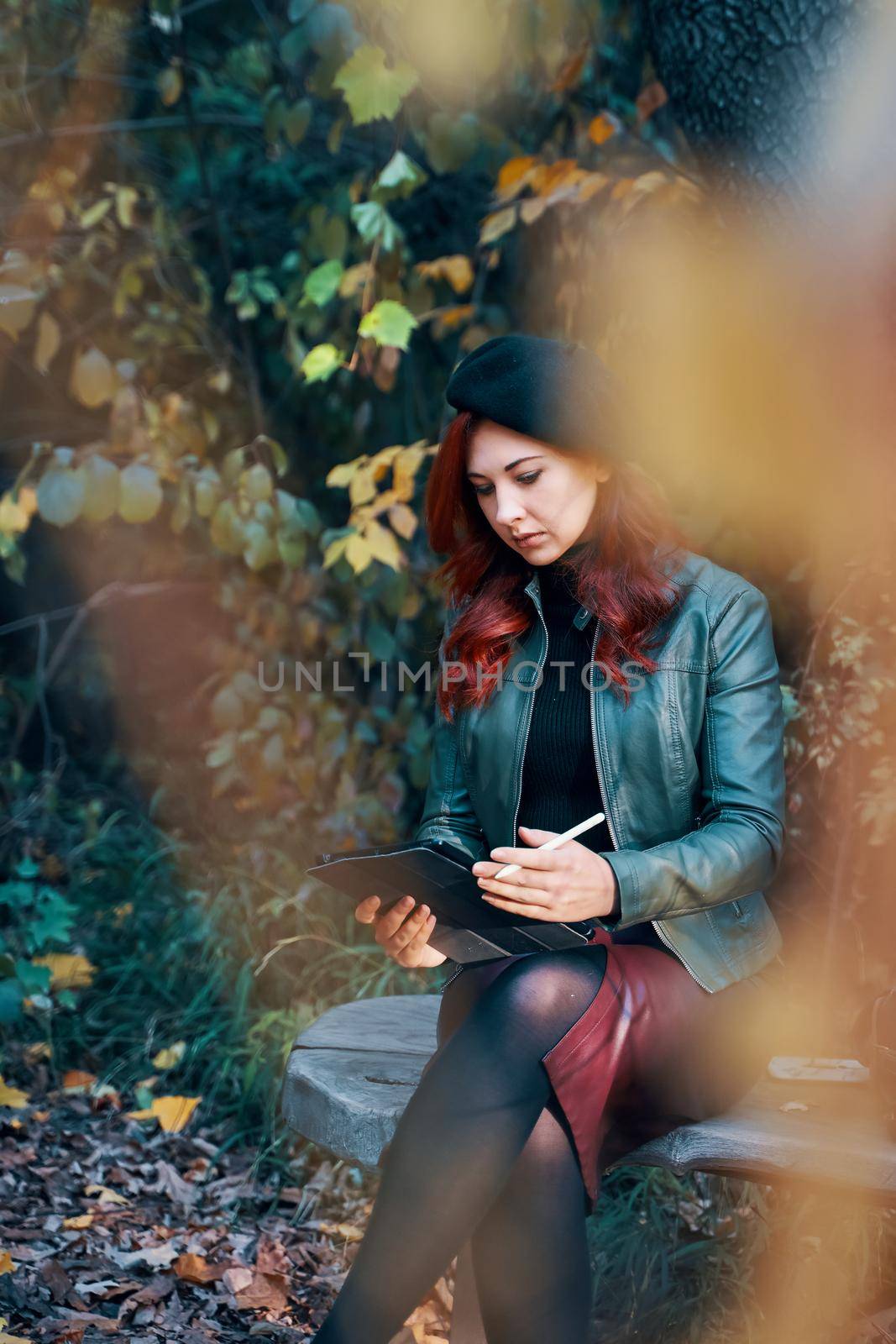 Red-haired girl in spring park on bench. Beautiful woman in beret and leather jacket. Person with graphic tablet, designer, freelancer.