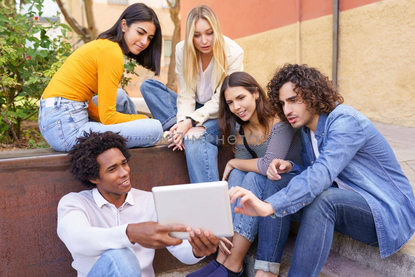 Young black man showing something on his digital tablet to his group of friends sitting on some steps in the street.