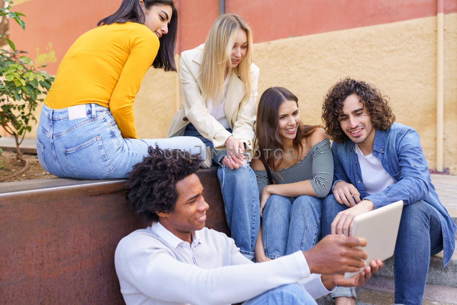 Multi-ethnic group of young people looking at a digital tablet outdoors in urban background. Group of men and woman sitting together on steps.