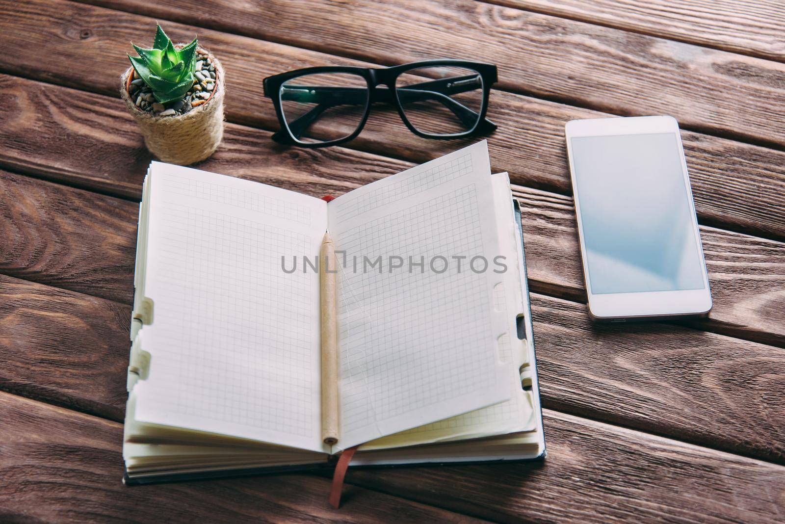 Notepad with pen, glasses and smartphone near a plant on a wooden table. Business working place.