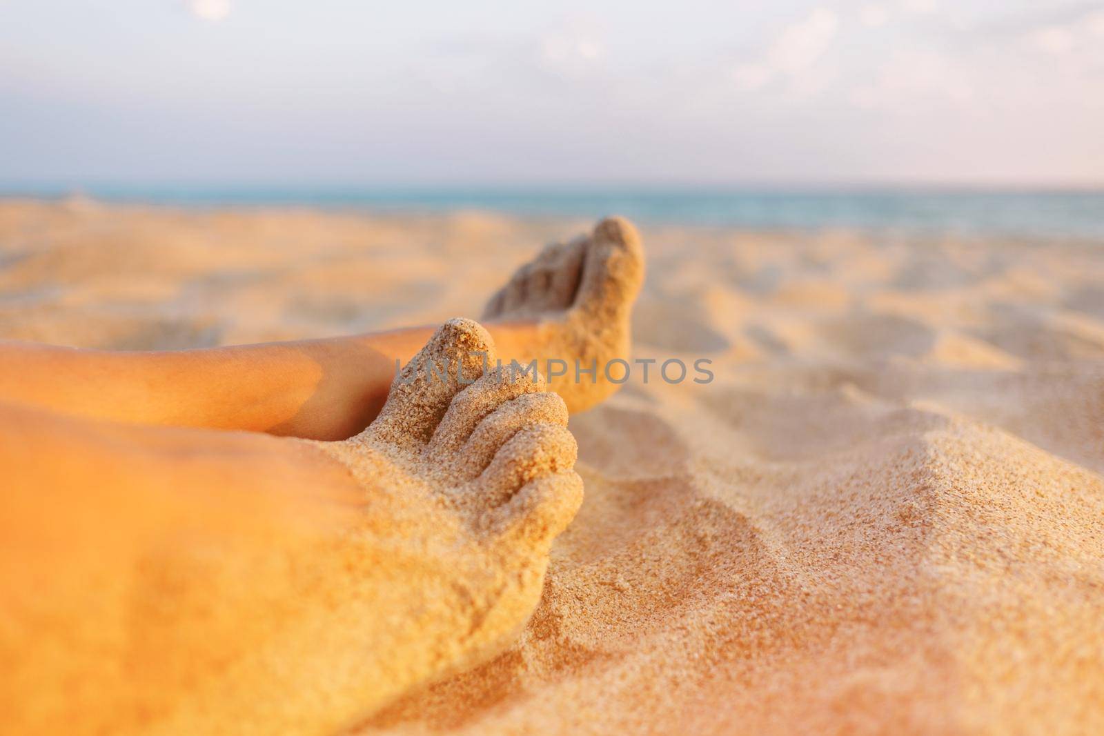 Woman resting on beach near the sea, close-up view of female barefoot legs on sand.