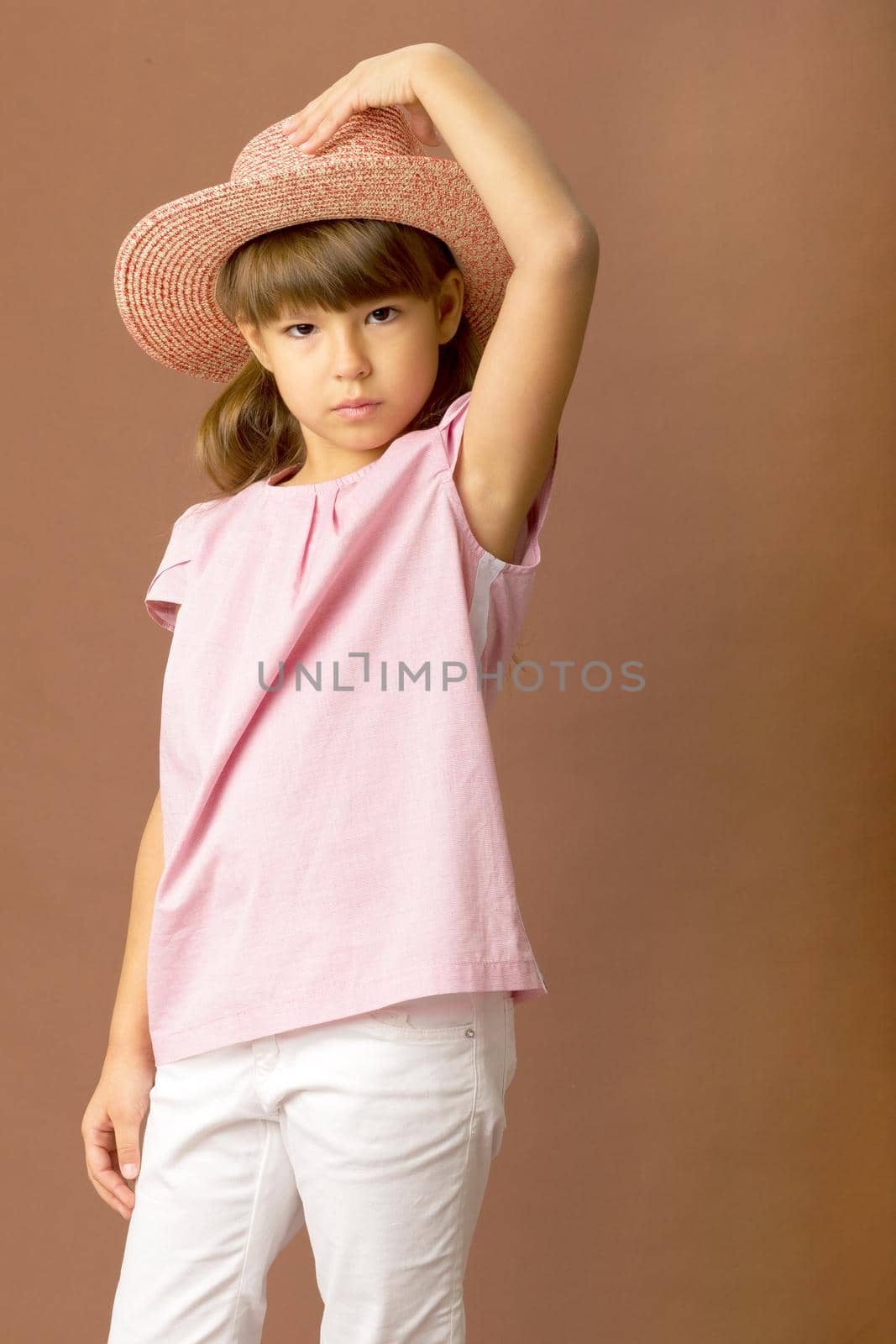 Adorable girl touching her straw hat. Child in pink t-shirt, white pants and straw hat posing on brown background in studio. Full length portrait of cute preteen girl looking seriously at camera