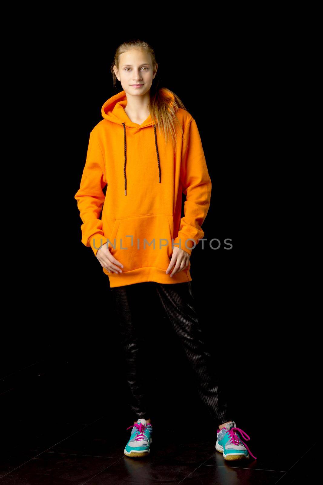 Full length shot of stylish teenage girl. Joyful girl with ponytail wearing oversized orange hoodie and black leather pants standing against black background in studio