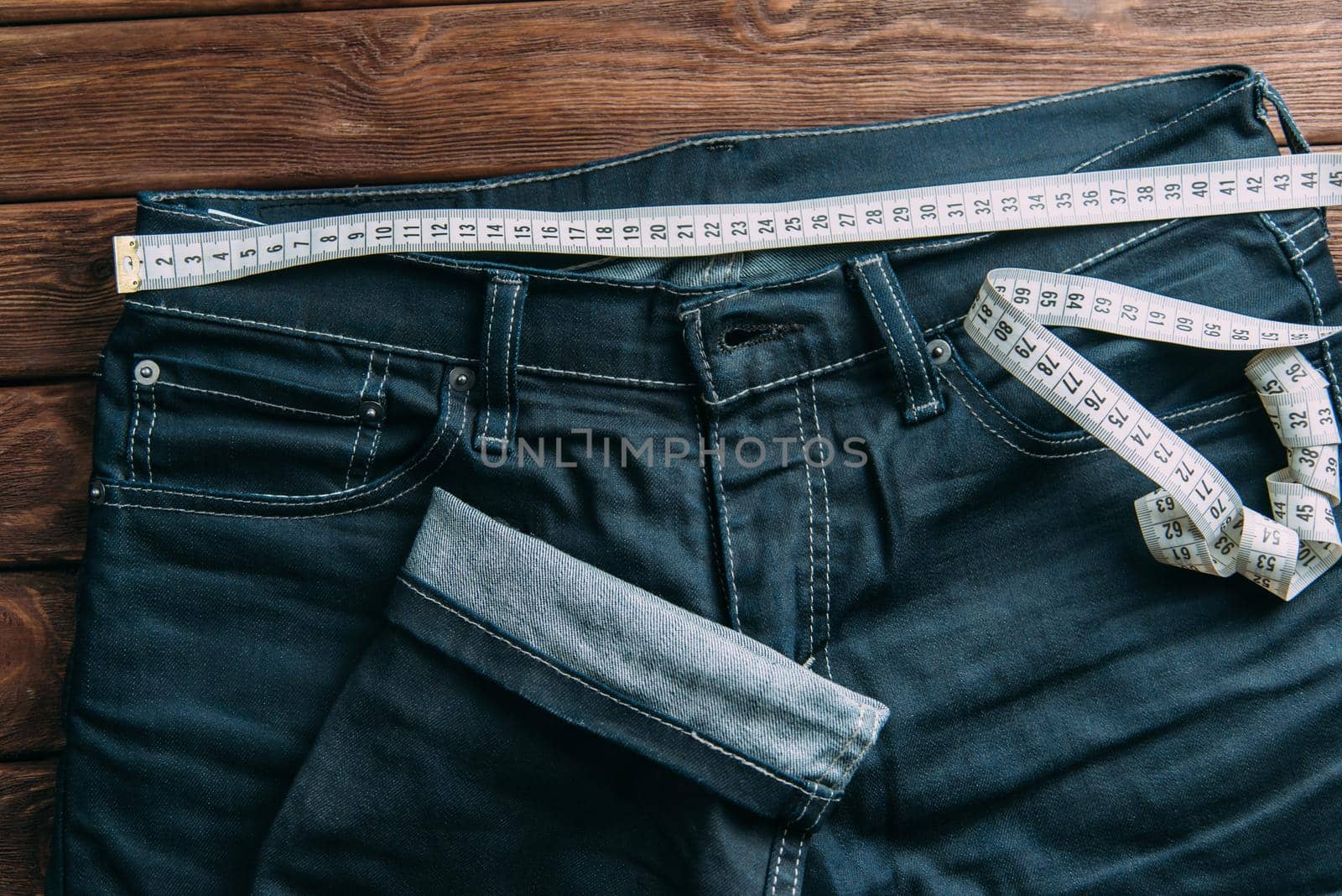 Jeans pants and measuring tape on a wooden background, top view.