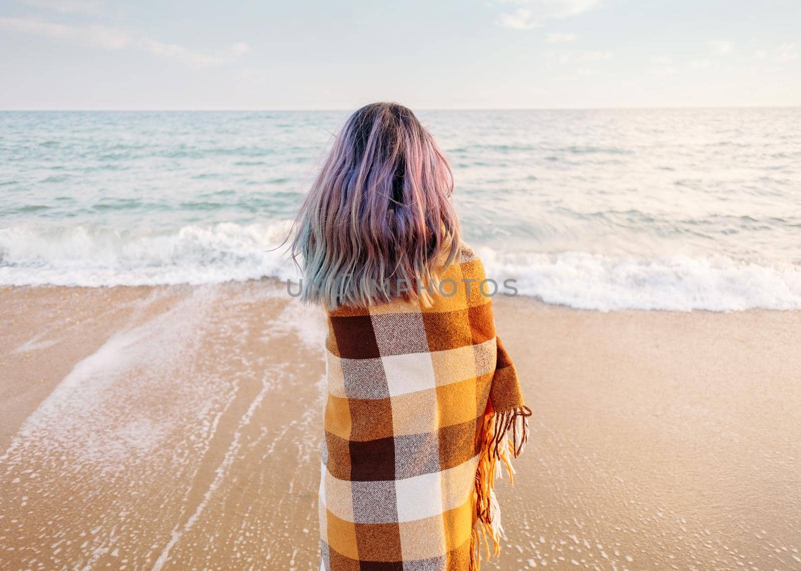 Young woman wrapped in a plaid standing on sand beach and looking at sea, rear view. Summer vacations.
