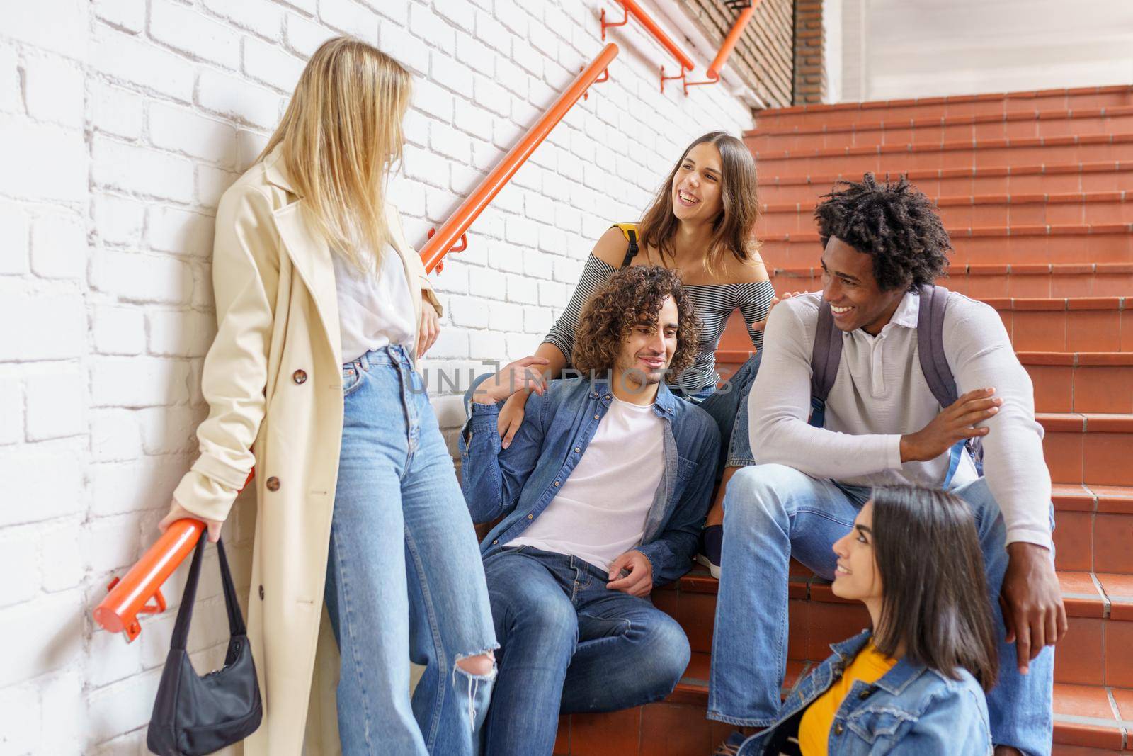 Group of friends with ethnic variety, sitting on some street steps having fun together outdoors.