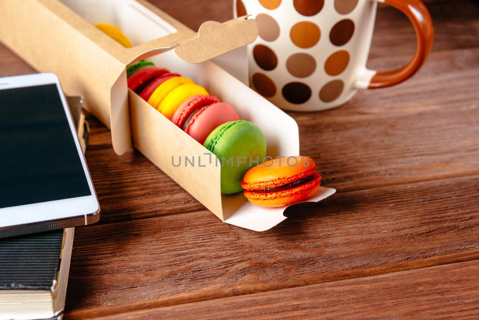 Sweet macarons and cup of drink on a wooden table near the smartphone and notepad.