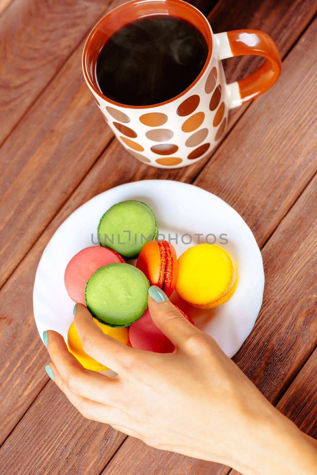 Female hand holding macarons cookie near the cup of hot drink on wooden table, top view.