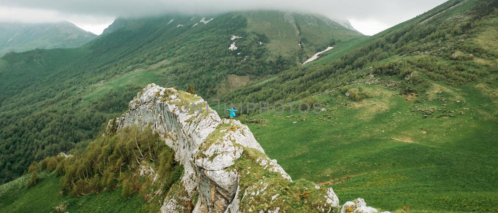 Small figure of traveler in raincoat standing with raised arms on cliff wall among mountains in summer in rainy weather.