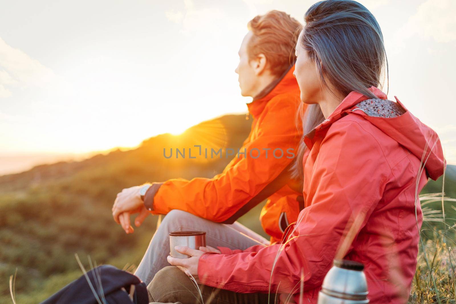 Traveler young couple resting with cup of hot drink in summer mountains and enjoying view of sunset light.