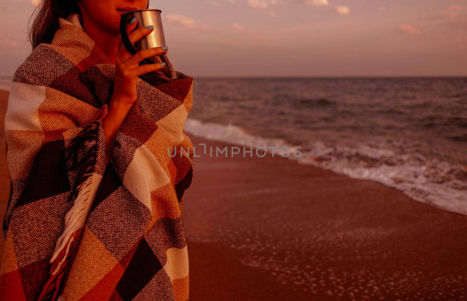 Young woman wrapped in plaid with cup of drink resting on beach on sunset.