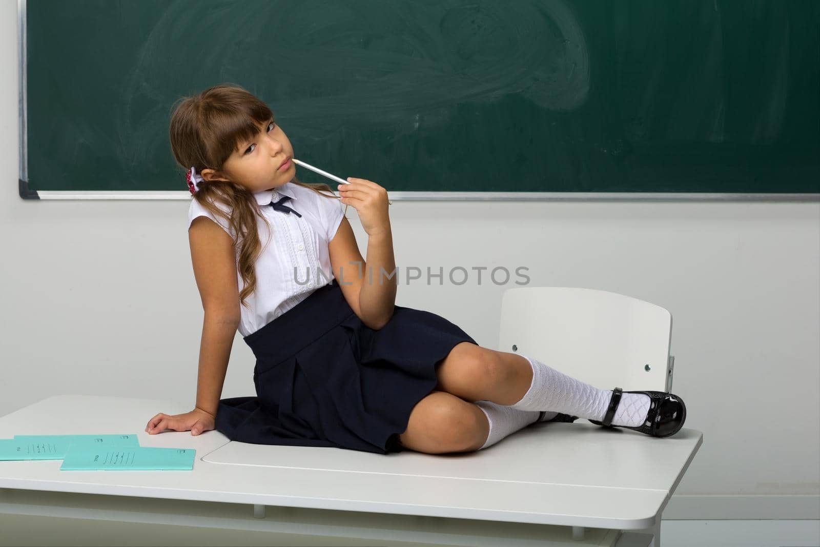 Cute girl sitting on desk in classroom. Adorable girl student in white blouse and blue skirt holding pen in her hand posing at blackboard. School and education concept