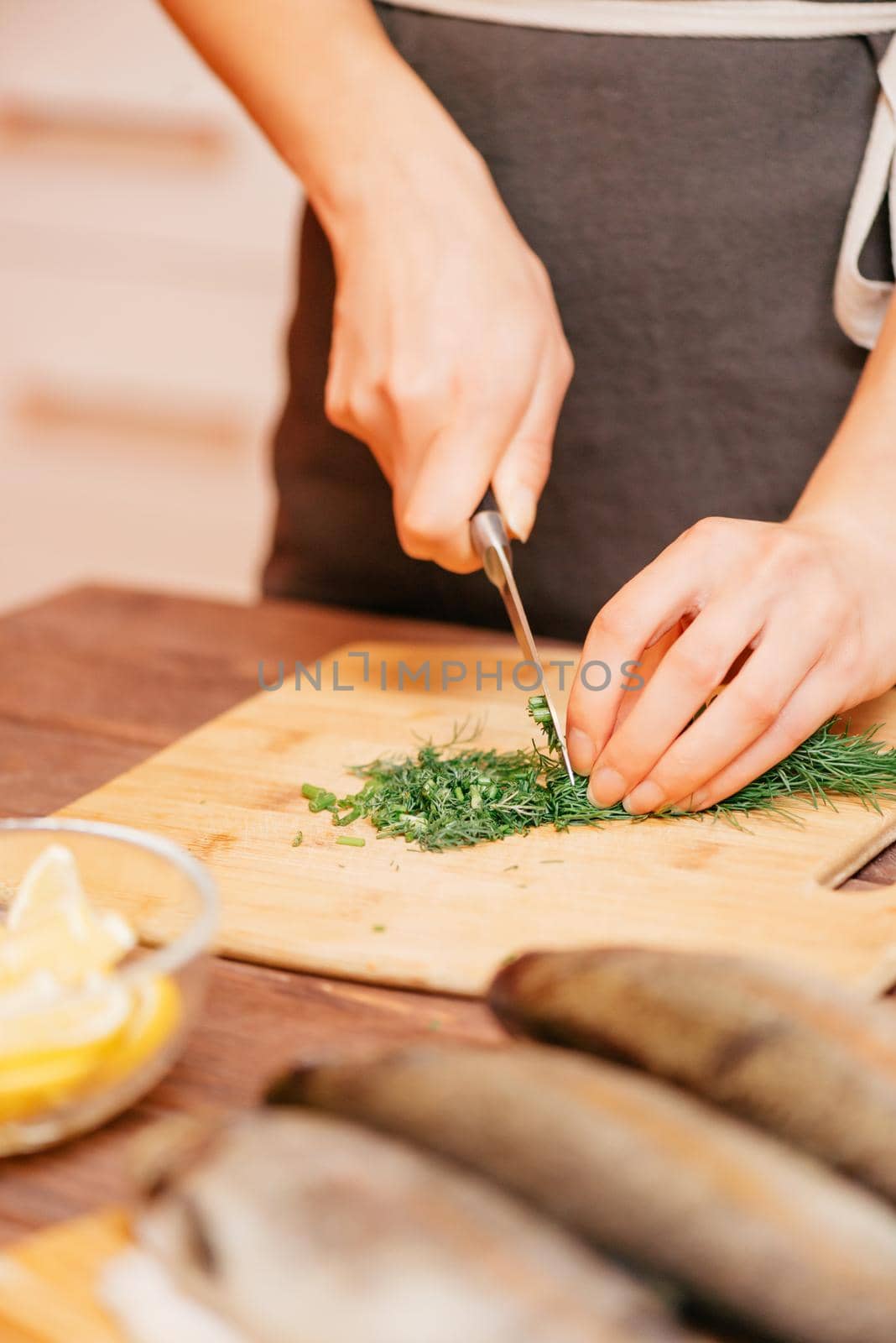 Woman cutting greenery on wooden board in the kitchen for fish dish, view of hands.