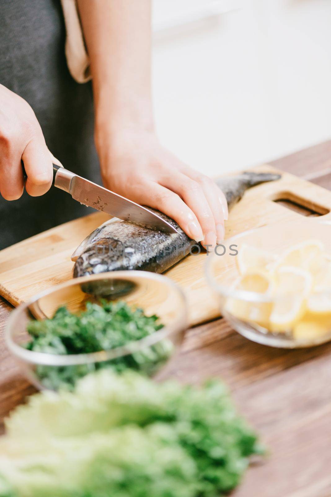 Unrecognizable woman cutting raw fish on wooden board for dish in the kitchen, view of hands.