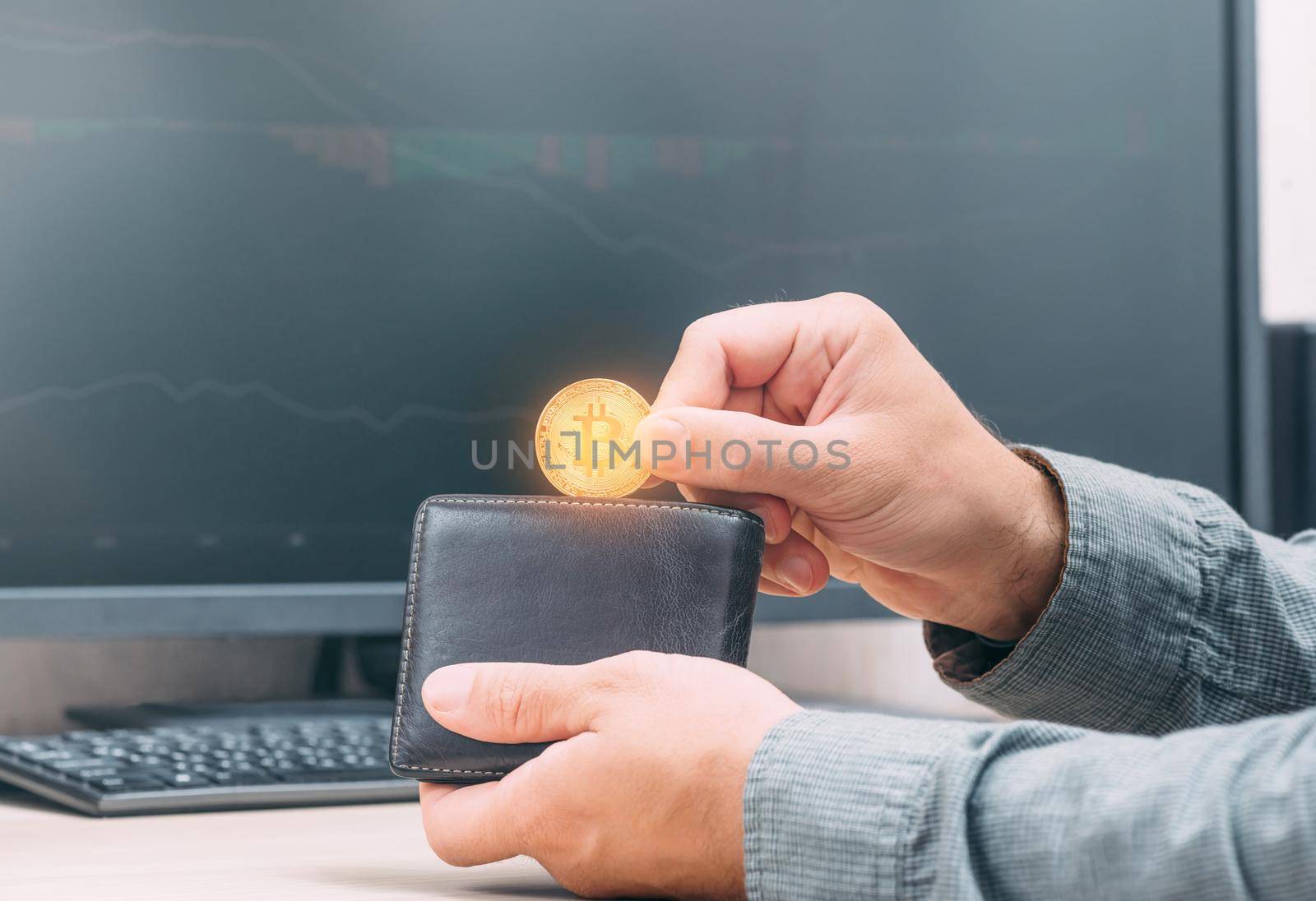 Unrecognizable man holding gold bitcoin and putting into wallet on background of infographics on computer screen.