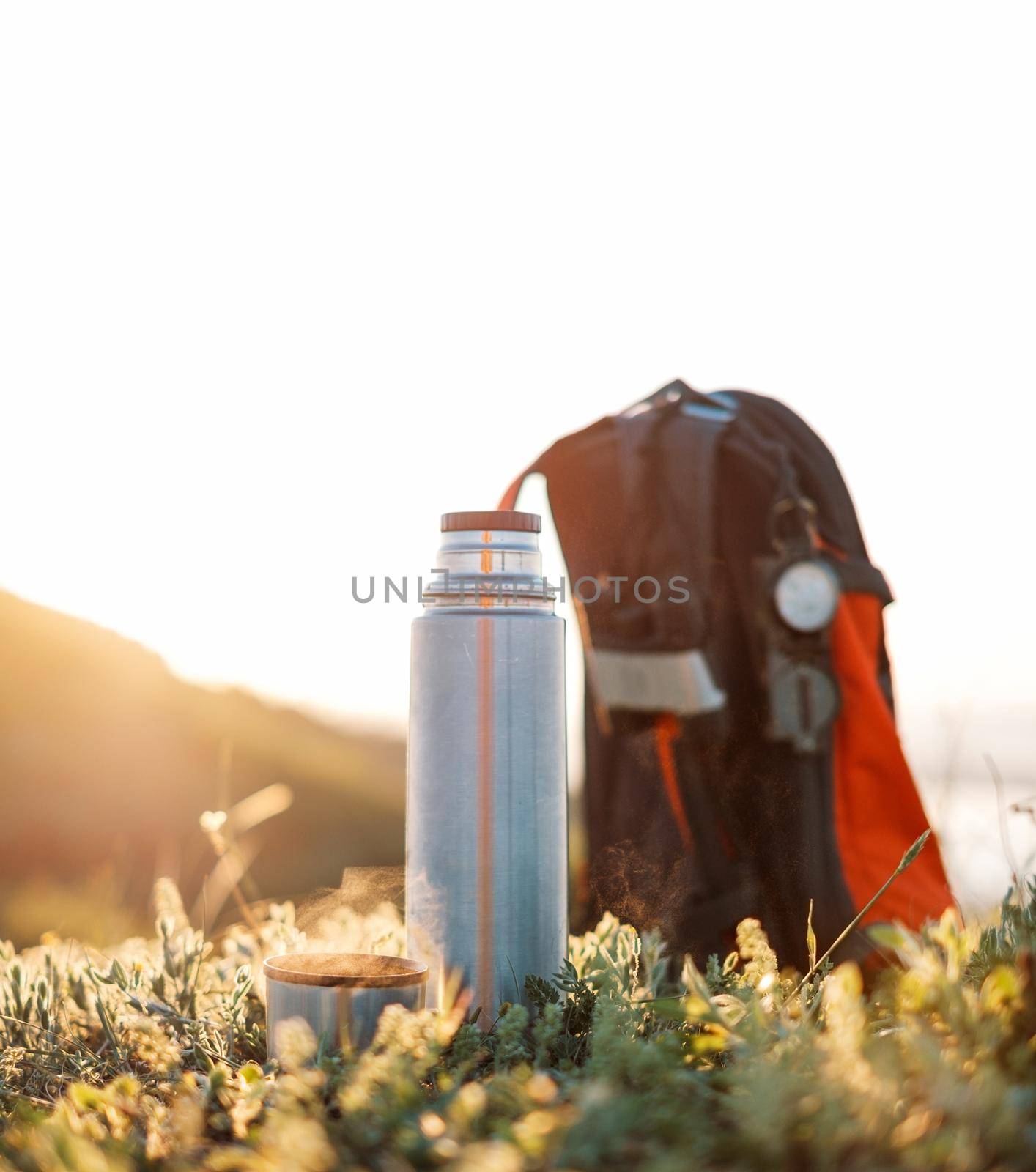 Thermos and cup of hot beverage near the backpack on green grass outdoor in summer.