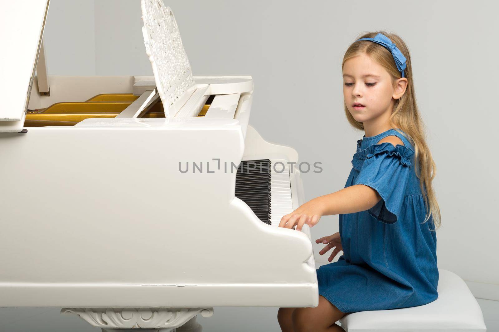 Charming girl posing at grand piano. Portrait of smiling six or seven years old girl posing at musical instrument. Portrait of adorable child wearing denim dress looking cheerfully at camera