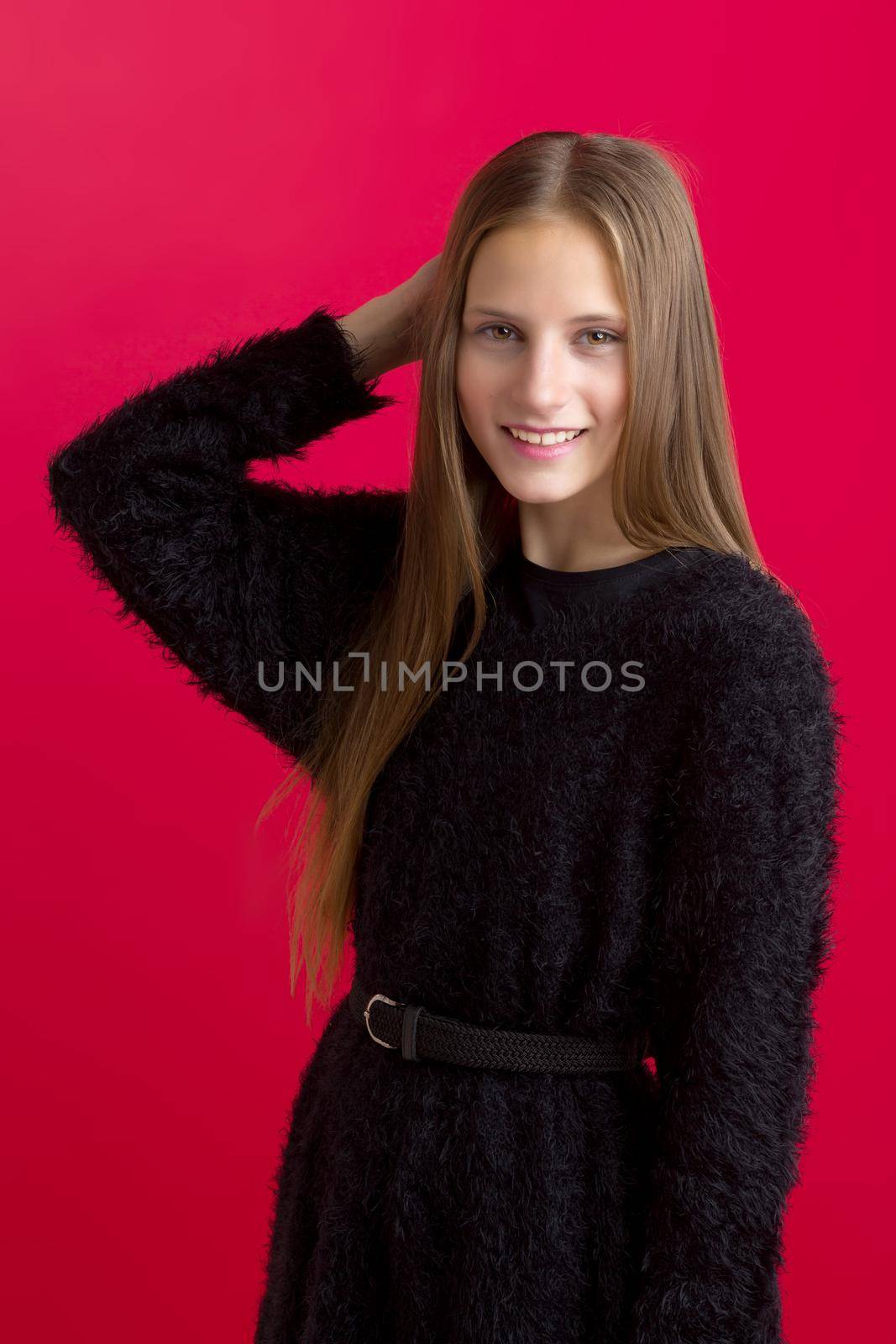 Beautiful teenage girl touching her long hair. Portrait of charming smiling teenager wearing black stylish oversized sweater with leather belt standing on red background