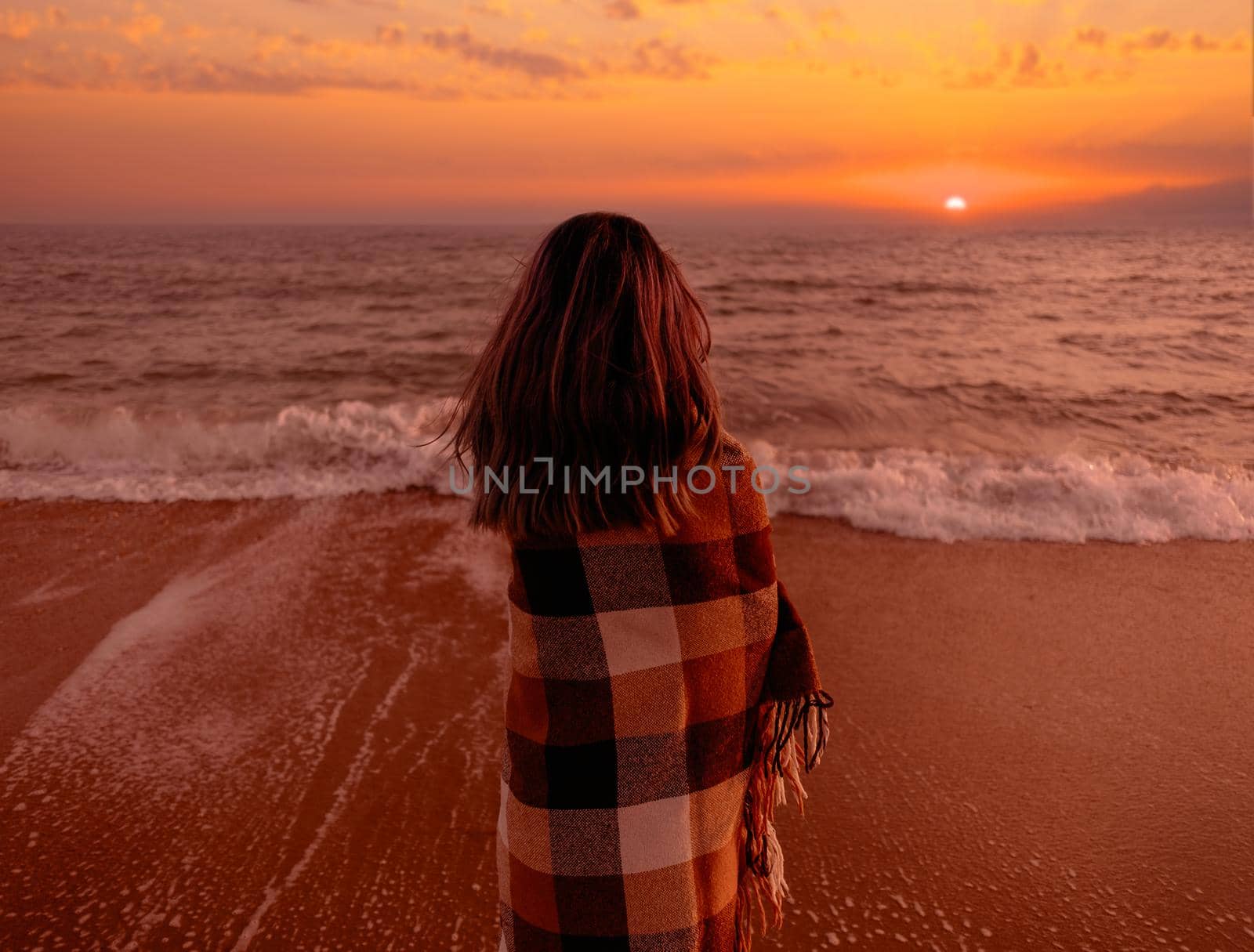 Young woman wrapped in a plaid standing on sand beach and looking at sea at sunset. Summer vacations.