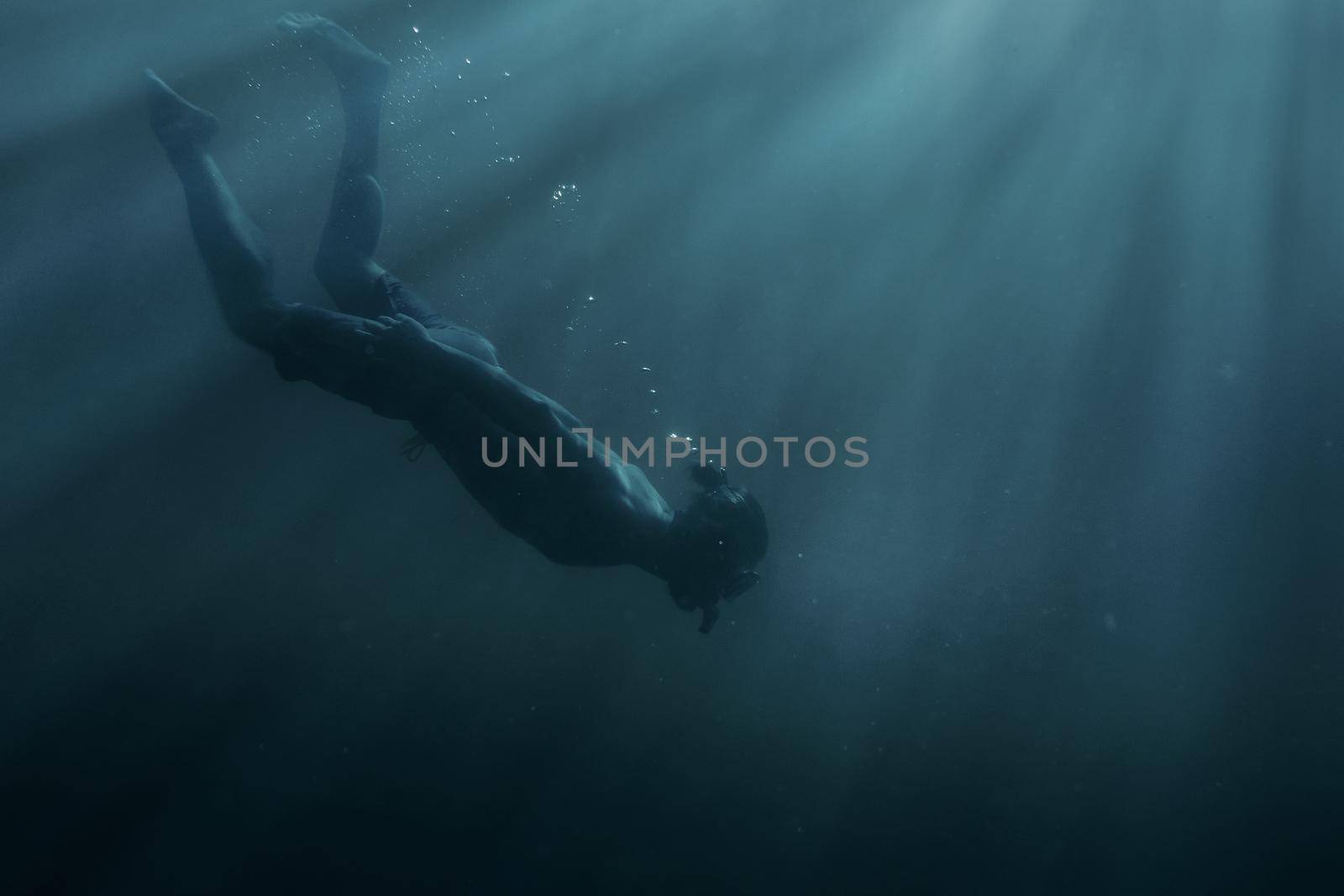 Young man freediver swimming with mask and snorkel underwater in deep sea among sunbeams.