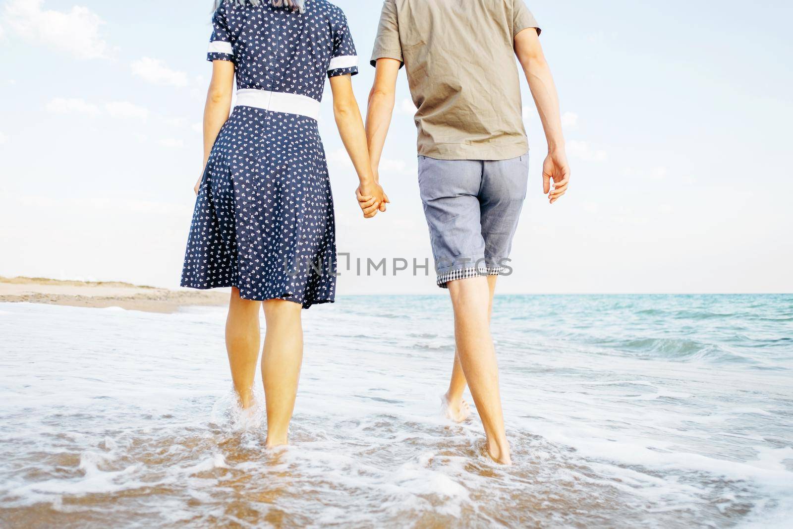 Unrecognizable young couple in love walking on sea coast and holding hands, summer vacations, rear view.