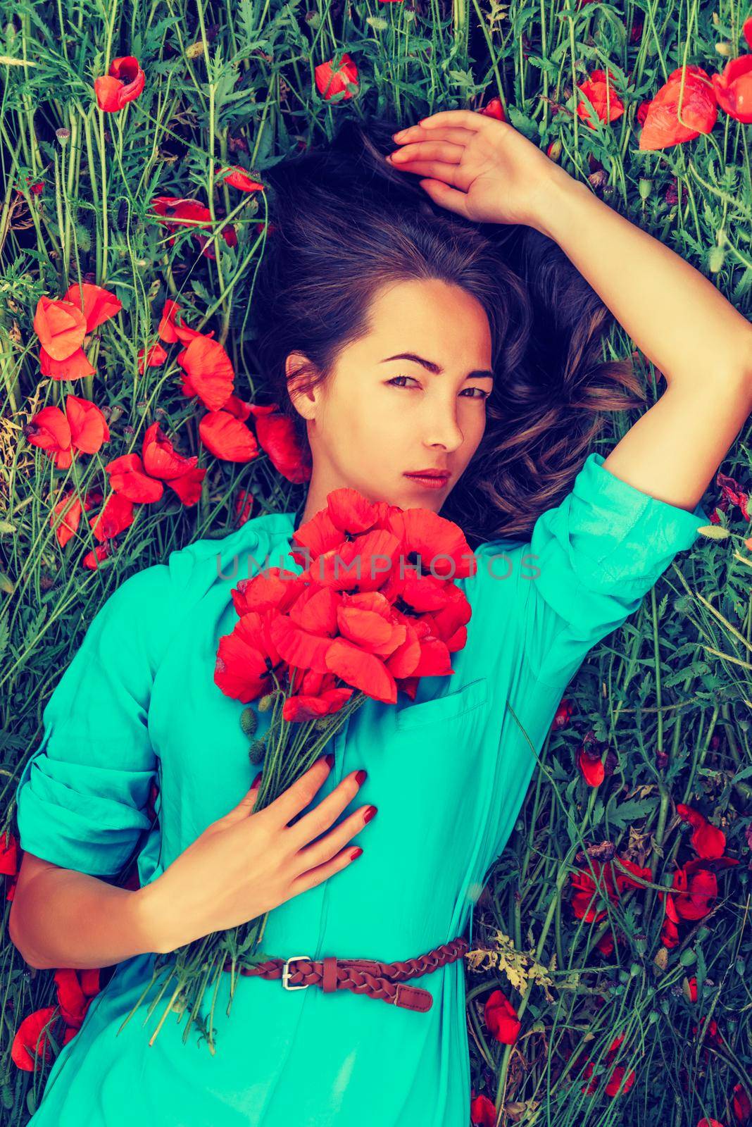 Beautiful young woman lying on red poppy meadow with bouquet of flowers, looking at camera. Beauty and fashion concept. Toned image.