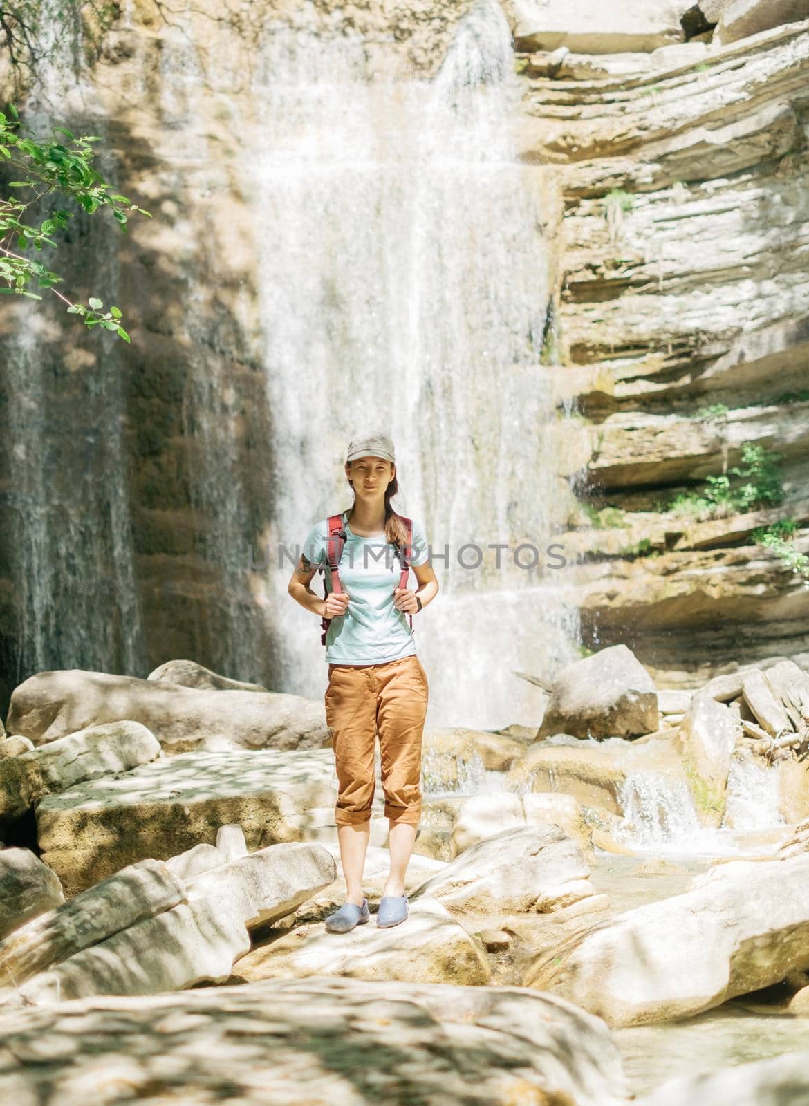 Traveler young woman with backpack standing on background of waterfall and looking at camera on sunny summer day.