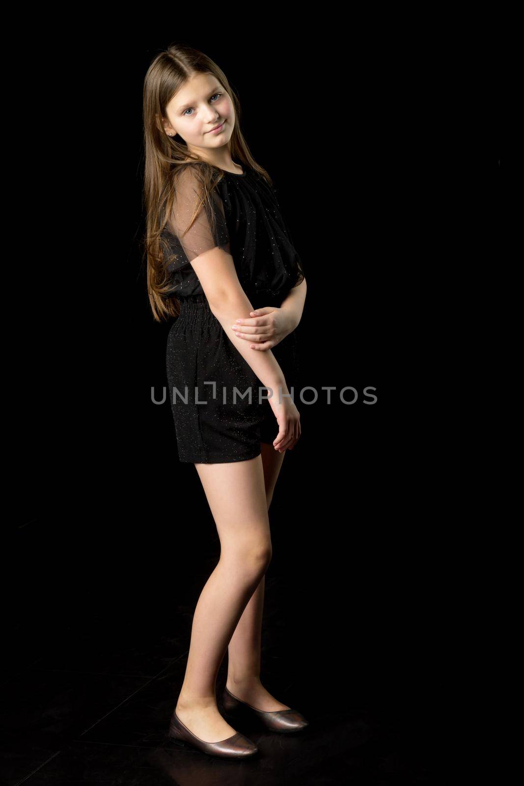 Full length shot adorable girl smiling at camera. Cheerful long haired teenage girl dressed in black jumpsuit and shoes standing against black background in studio and staring at camera