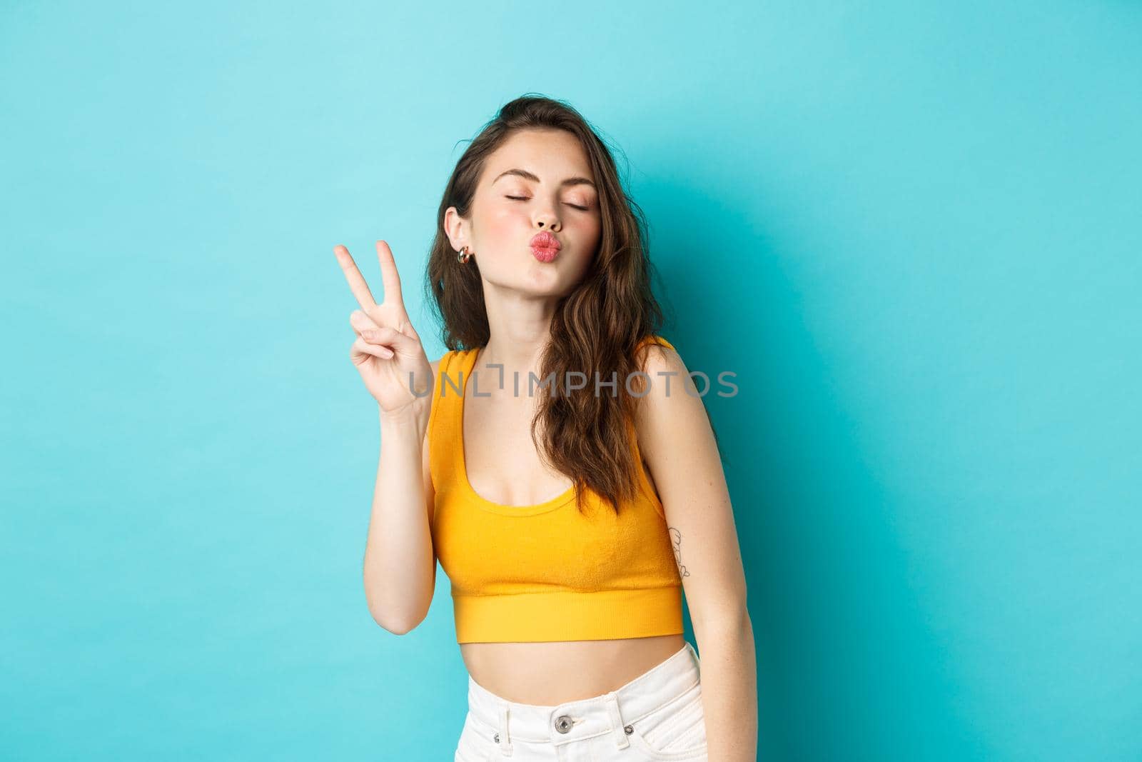 Glamour girl in summer clothes close her eyes and making kissing face with v-sign, enjoying vacation, having fun, standing relaxed against blue background.