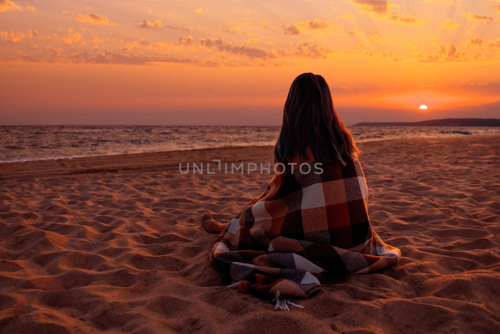 Unrecognizable woman wrapped in plaid resting on sand beach and enjoying view of sunset over sea.