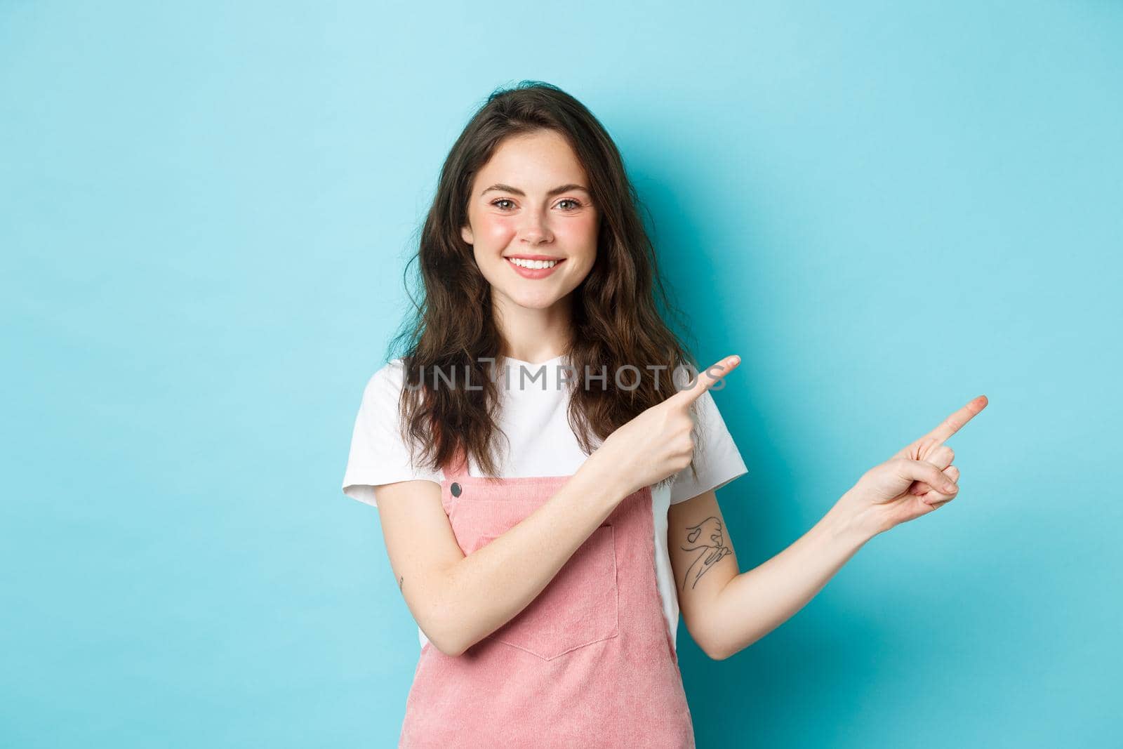 Image of cute teenage girl smiling, pointing fingers right at promo offer, demonstrating banner on copy space, standing in summer clothes with glamour make-up against blue background by Benzoix