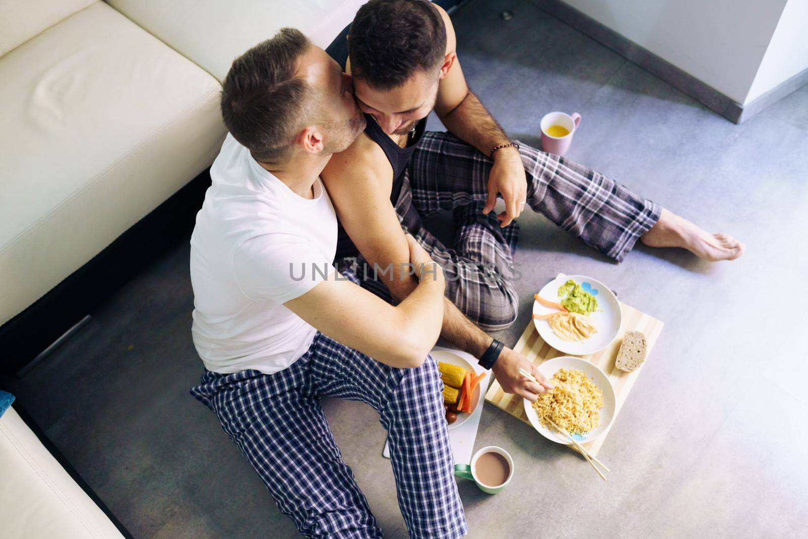 Gay couple eating together sitting on their living room floor. Homosexual lifestyle concept.