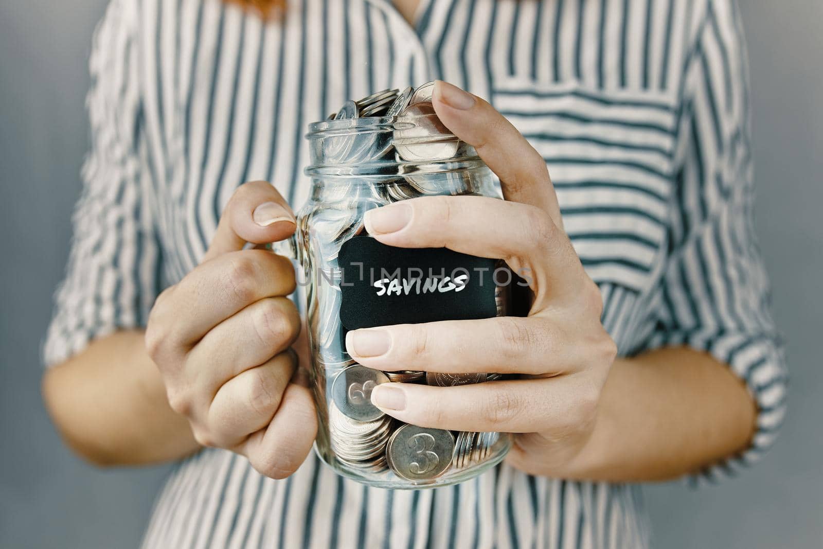Girl n white and black striped shirt holds her savings. Glass bottle with silver coins in women's hands. Sticker with inscription on money jar.