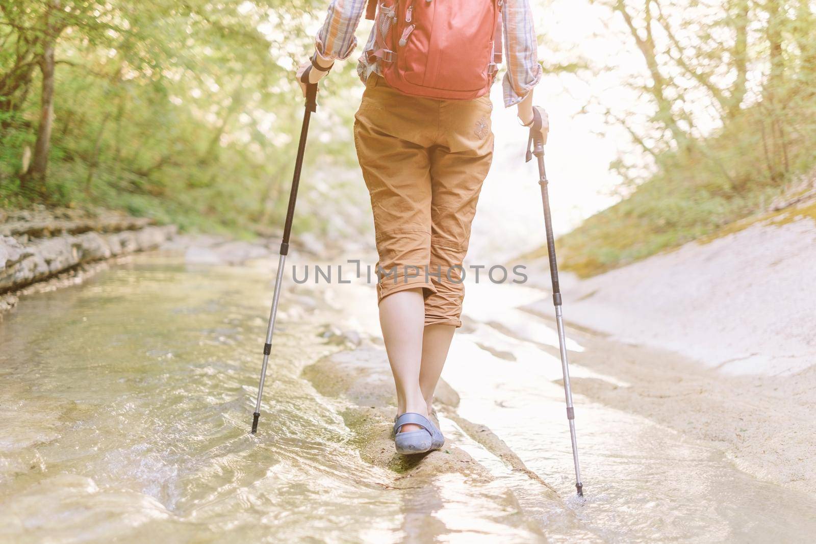 Traveler backpacker woman with trekking poles walking along the river in summer outdoor, view of legs.