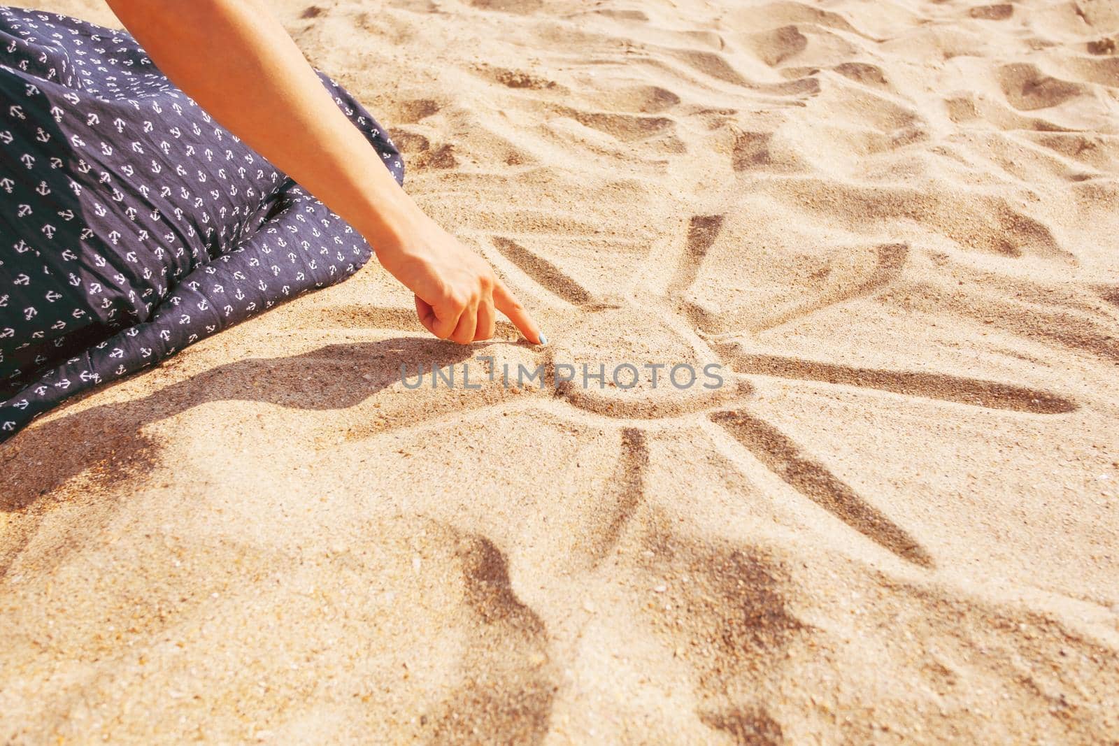 Unrecognizable girl draws a sun with sunbeams on sand beach with her finger, view of hand.